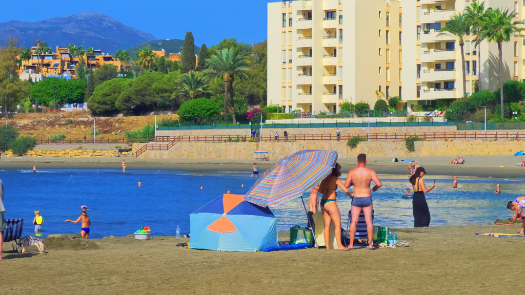 Beachgoers beside a striped parasol at Playa del Cristo, with apartments behind the bay.