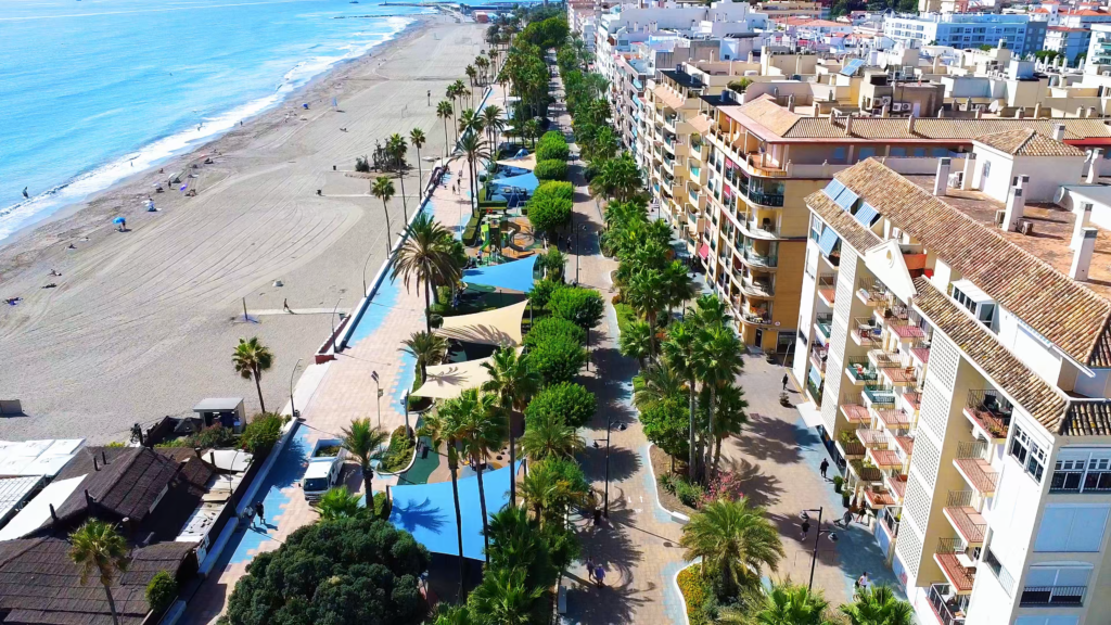 Aerial view along Estepona’s promenade with palm trees, shaded play areas and beachfront apartments.