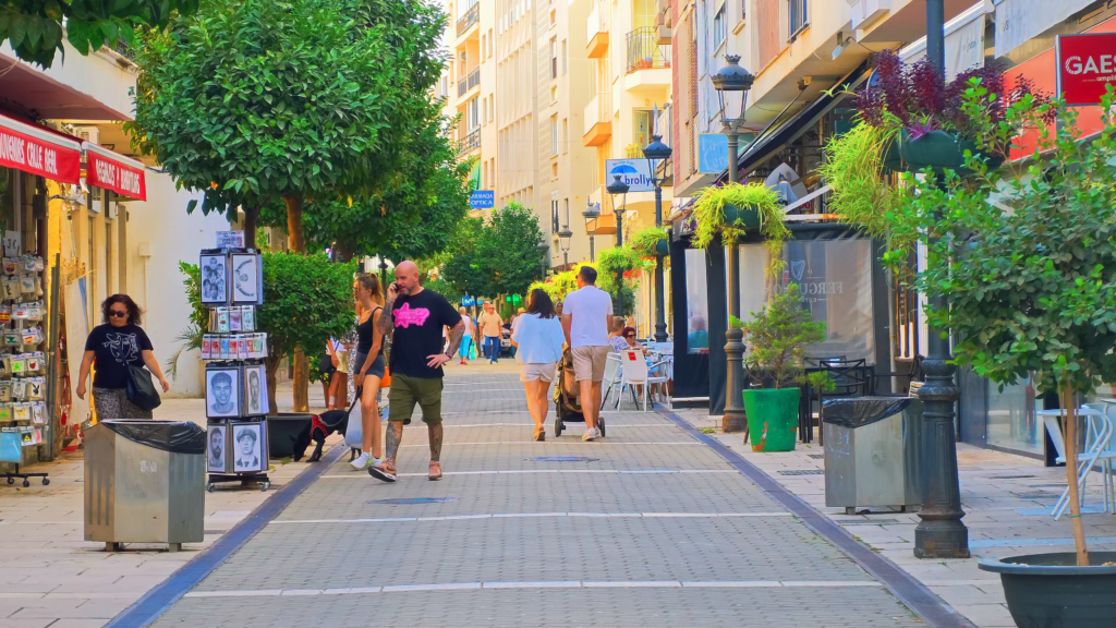 Pedestrians walking along a shopping street in Estepona lined with cafés, trees and flowers.